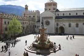 Una piazza ampia e assolata è animata da diverse persone che passeggiano attorno a una fontana elaborata, con una maestosa cattedrale e un complesso di antichi edifici sullo sfondo di montagne.