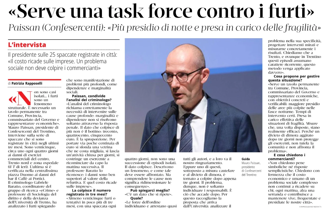 Al centro, un uomo con occhiali e completo parla a un microfono, affiancato da un articolo di giornale stampato.