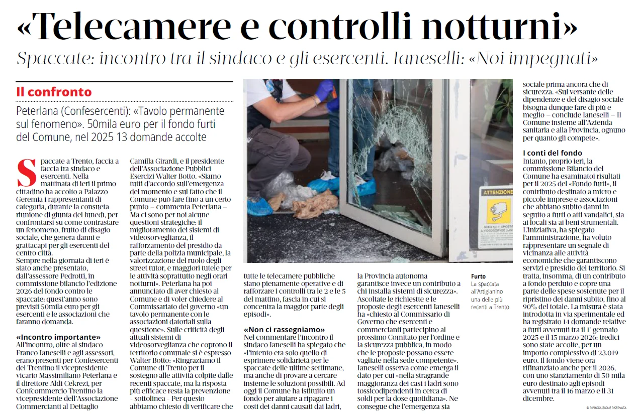 Una persona con copriscarpe blu è chinata accanto a una porta di vetro in frantumi, con frammenti di vetro sparsi sul pavimento, alcuni detriti e un cartello di avviso giallo con simbolo di telecamera visibile sulla destra.